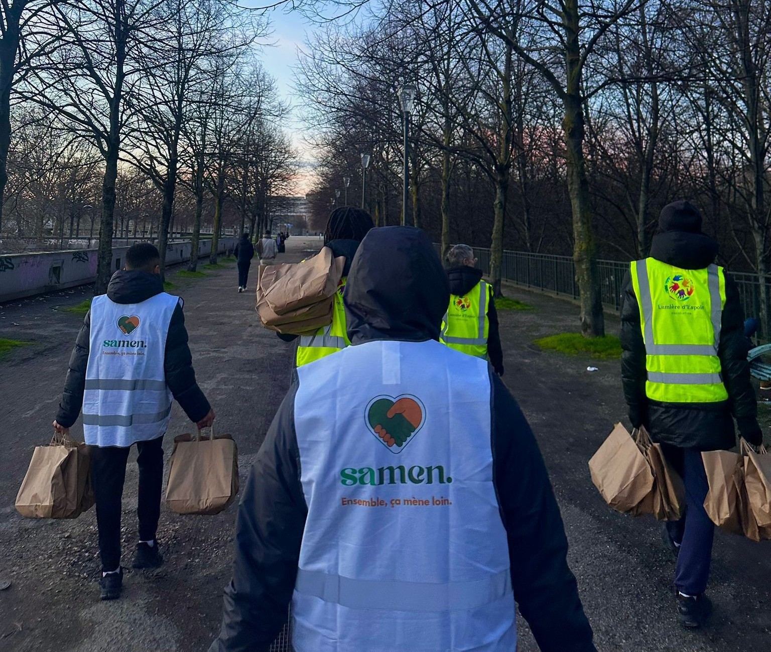 Des bénévoles de Samen partent en maraude, sacs de repas à la main, le long d'une allée d'arbres au crépuscule.