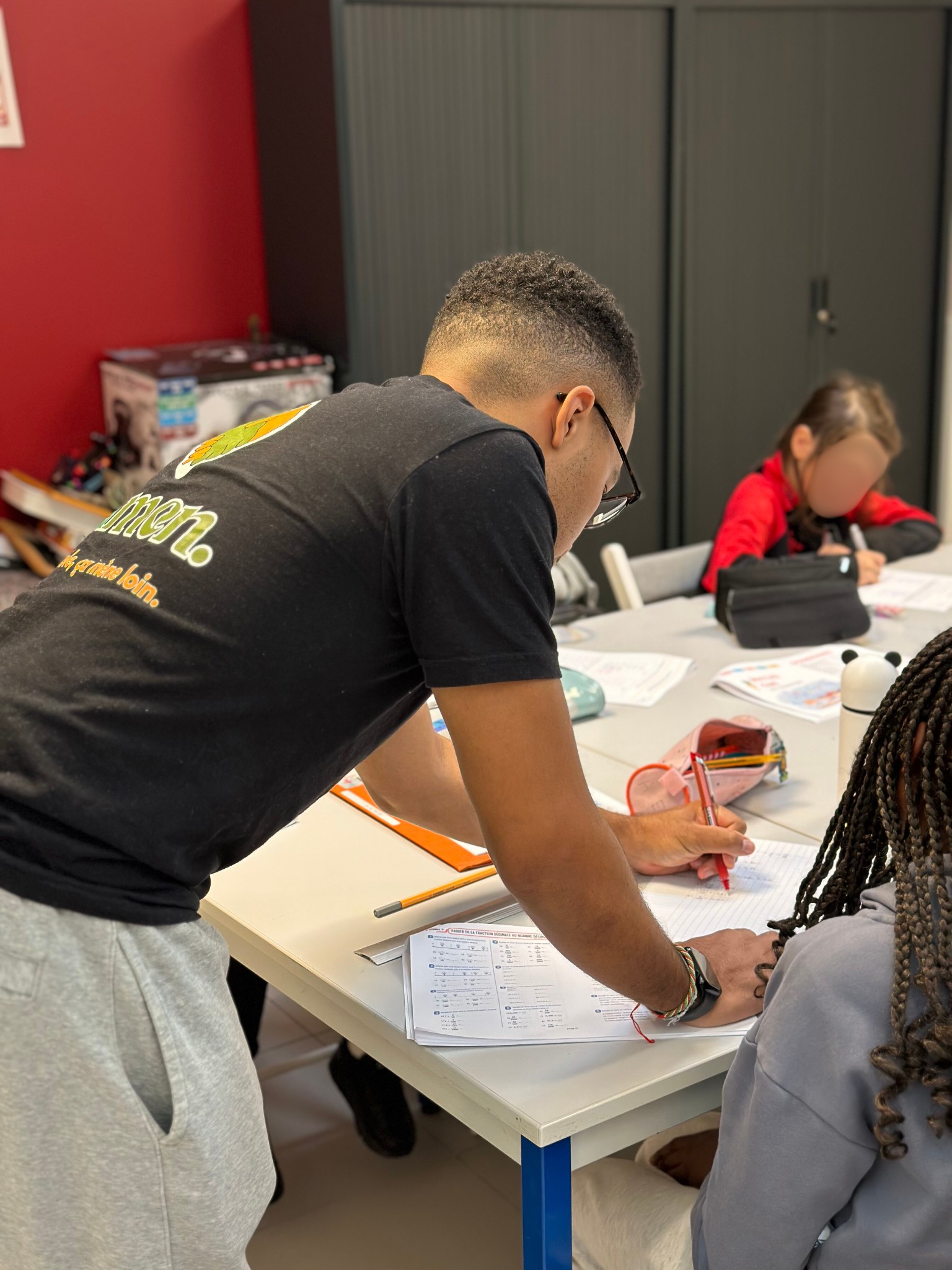 Un bénévole de Samen aide deux enfants à faire leurs devoirs autour d'une table de travail.